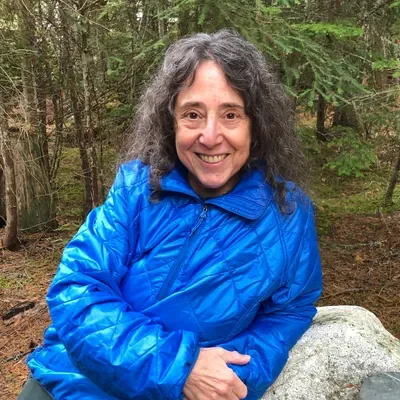 Photo of Esta Porter, a woman with wavy grey hair, smiling into the camera. She is wearing a blue rain jacket, and leaning on a rock in the rainforest.